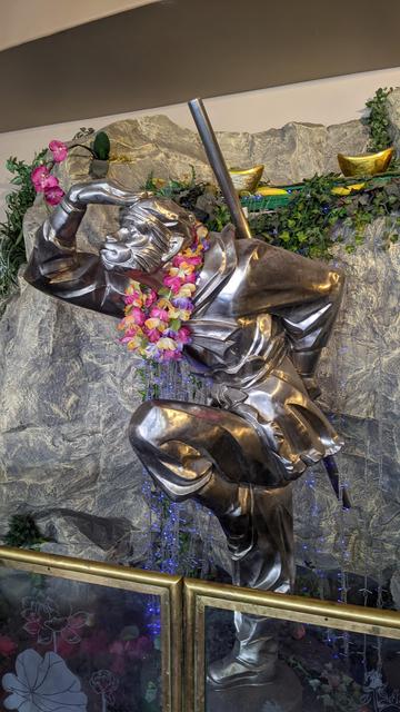 A silver statue of Sun Wukong, the Monkey King, from the story "Journey to the West." He is standing with his right hand perpendicular to his forehead as if looking out off into the distance and shading his eye from the sun. With posture leaning forward. His left hand is holding his magical staff, the Ruyi Bang. His left leg is bent at the knee, raised up, with his other foot firmly planted on the ground.