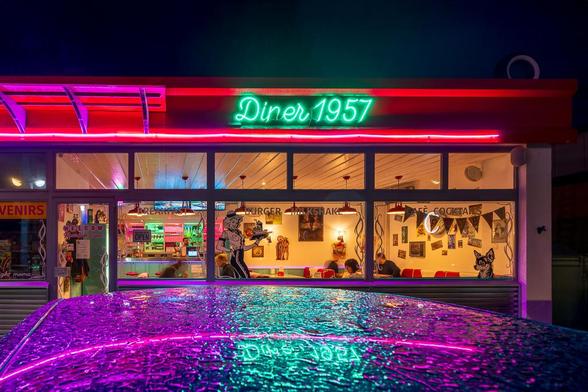 Im Vordergrund das Dach eines Autos, nass vom Regen. In den Regentropfen, spiegelt sich das Diner im Hintergrund.
Man sieht die Fensterfront, erkennt einige Kunden im Diner. Das rote Dach ziehrt einen grüner Neonschriftzug : Diner1957.