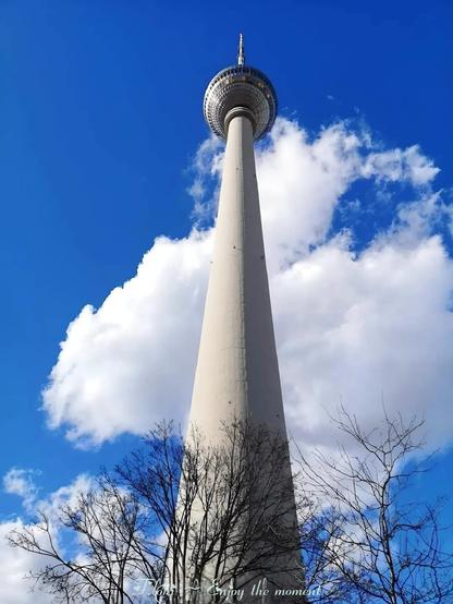 Das Bild zeigt den Berliner Fernsehturm, der imposant in den strahlend blauen Himmel mit einigen weißen Wolken ragt. Die Aufnahme wurde aus der Froschperspektive gemacht, wodurch der Turm besonders hoch und beeindruckend wirkt. Im Vordergrund sind einige kahle Baumzweige zu sehen, die einen Kontrast zur massiven Struktur des Turms bilden. Am unteren Rand des Bildes steht in weißer, geschwungener Schrift der Text: "F.loki ~ Enjoy the moment".