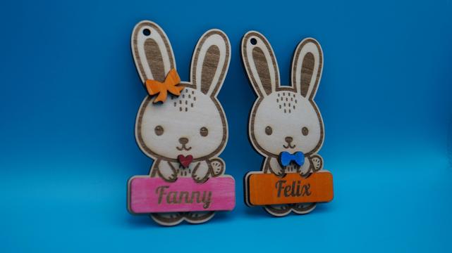 Zwei liebevoll gestaltete Osteranhänger aus Holz in Hasenform mit Gravur – links ein Hase mit rosa Namensschild "Fanny" und orangefarbener Schleife, rechts ein Hase mit orangem Namensschild "Felix" und blauer Fliege. Beide Anhänger sind handbemalt und vor einem blauen Hintergrund fotografiert.
