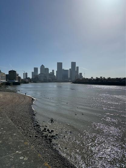 A view of the Canary Wharf skyline, taken further west along the Thames Path in Wapping. Sunshine is reflecting in sparkles on the river’s surface. Clear blue skies above the high-rise buildings. 