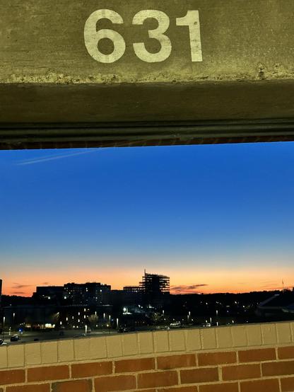 Color photo of a sunset against a city skyline framed at top and bottom by the inside of a parking garage.