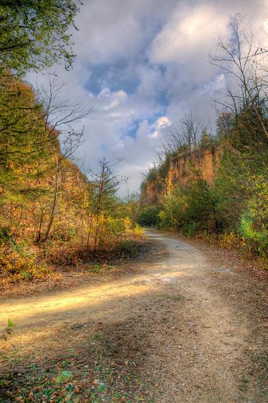 A winding dirt path cuts through a serene autumn landscape, framed by towering cliffs and vibrant foliage.