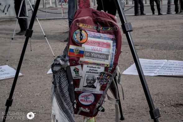 Ein Rucksack, der unter einem Kamera-Stativ hin. Pro-Russische Patches, Julian Assange, Friedensbewegung und Palästina sind vertreten. Palästinasolidarität wird mit der Samidoun-Faust dargestellt.