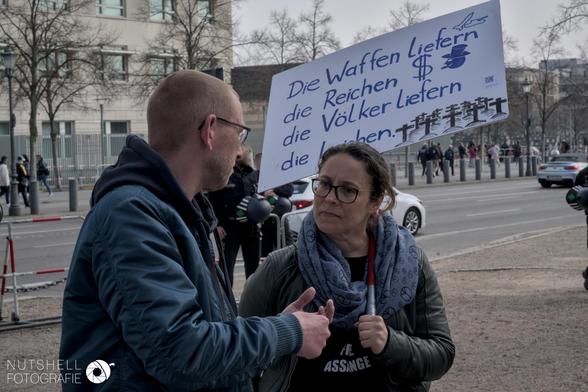 Ein Schild mit der Inschrift "Die Waffen liefern die Reichen $ <smiley mit Zylinder> die Völker liefern die Leichen <Kreuze>". In Hintergrund das Holocaust-Mahnmal zu sehen.