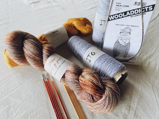 Four skeins of wool in yellow, light lavender and speckled brown with few needles in front on a white table cloth. 