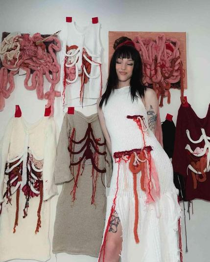 Woman wearing knitted garments adorned with representations of internal organs and blood, stands in front of similar works displayed on a wall.