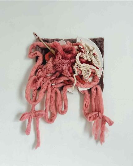 Abstract wall hanging of tangled, pink and red dyed wool threads representing intestines in a shallow white dish with a wooden spoon.