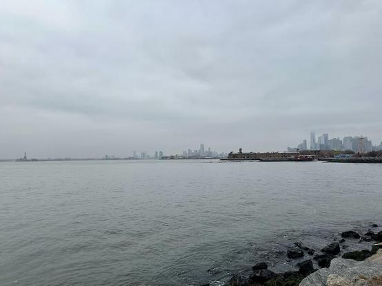 Photo of New York Harbor on a sad, gray, cloudy day, looking northwest from somewhere in Red Hook. The Statue of Liberty is at far left. The towers of downtown Manhattan are close to far right. Not much chop on the water, though.