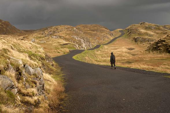 Une route étroite serpente du premier plan vers le haut de l’image, à travers un paysage accidenté couvert d’herbes jaune. Le ciel est sombre, une personne, de dos, marche vers l’horizon. 