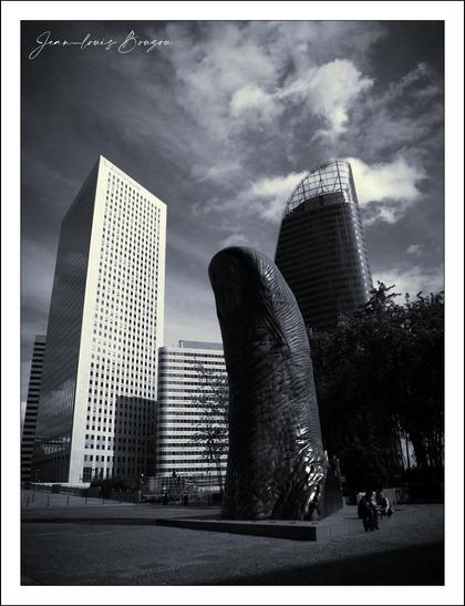 The image captures a striking urban scene, prominently featuring a large, abstract sculpture of a thumb figure, standing amidst modern high-rise buildings. The sculpture, crafted with a glossy, dark material, contrasts sharply against the sleek glass facades of surrounding skyscrapers.
The setting is bathed in a monochromatic palette, lending the scene a dramatic quality. Wispy clouds in the sky add depth to the composition, creating a dynamic background against the stillness of the structures below. The juxtaposition of the organic shapes of the sculpture with the rigid lines of the buildings highlights the intersection of art and architecture in the urban landscape.
Symbolically, the sculpture may represent themes of progress and human endeavor, while the surrounding buildings signify innovation and the modern city life. The overall composition invites viewers to reflect on the relationship between nature and human-made environments, as well as the interpretations of dominance and strength conveyed through the raised form of the sculpture.