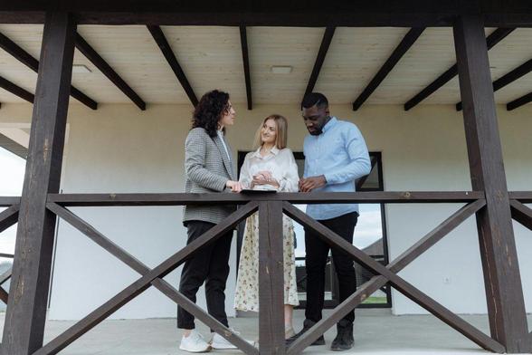 Three individuals standing on a porch in front of a house, smiling and engaging with each other