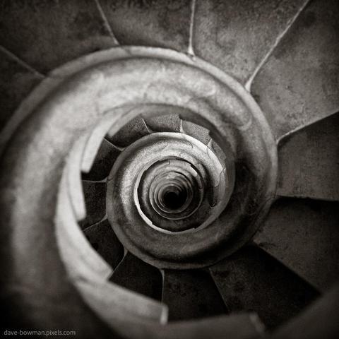 A photograph of a spiral staircase inside one of the Sagrada Familia steeples in Barcelona, Spain, shows its elegant, winding descent. Taken from above, the image highlights the staircase’s geometric patterns and intricate architectural details, a hallmark of Antoni Gaudí’s design. The spiralling steps narrow as they recede into the distance, creating a striking abstract composition in monochrome tones.