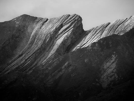 A monochrome image of the peak of the Sphinx Mountain in the Canadian Rockies. The clear sedimentary layers of the compressed rock can be seen, warped and tormented by the pressures of the earth as this mountain range formed. 
The late afternoon sun highlights the peaks adding extra contrast to the sharp layers.