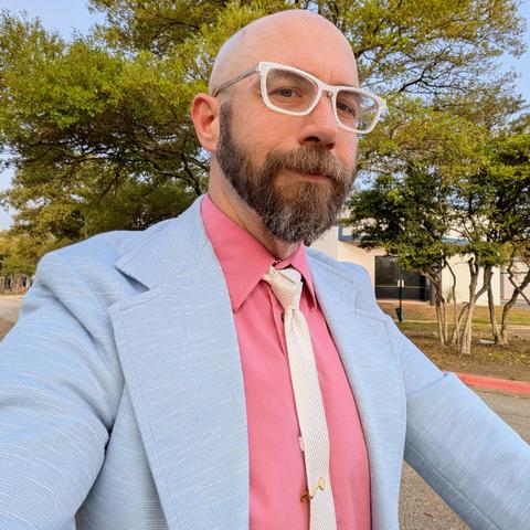 A photo of me, a 47 year old white man with a shaved head, a short cropped brown & grey beard, 

photographed from the chest up

In front of some live oak trees in an office park,

wearing

rectangular pearlescent white acetate glasses, a skinny white necktie in a trinity knot on a dusky pink dress shirt under a sky blue sport coat