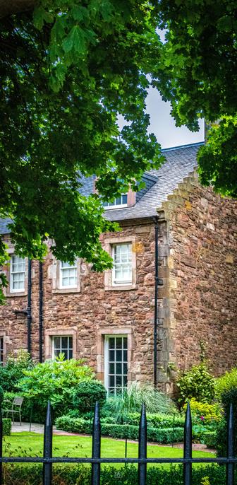 A quaint stone cottage partially hidden by trees and a spiked iron fence. Canongate, Edinburgh, 19 Aug 2024. Nikon D5600, Nikkor DX 35 mm ƒ1.8G, ISO 3200, ƒ13, -0.3 ev, 1/80s