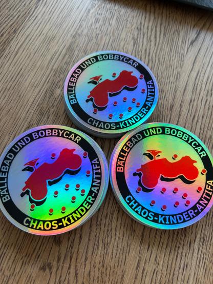 Bild zeigt 3 Stapel mit runden Stickern. In der Mitte ist die rote Silhouette eines BobbyCars zu sehen sowie rote Bälle. Auf einem schwarzen Band, das den Sticker umgibt, steht „Bällebad und Bobbycar - Chaos-Kinder-Antifa“  