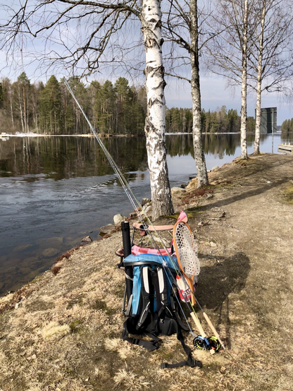 A camping chair, backpack and two fly rods on the river bank. The water surface is calm and the weather is sunny.