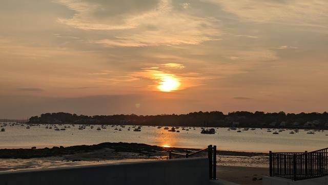Historical Documentation in the form of a photograph of a rising sun over the ocean bay at the Historic Pioneer Village in Forest River Park, Salem, MA. The sky is filled with orange and yellow hues, with the sun partially hidden behind the clouds giving them a shimmering silver look.  The sun is rising in the distance, casting a warm glow over the horizon. The water is calm and reflects the colors of the sky. In the foreground, there is a rocky shoreline with a few boats docked in the water. On the right side of the image, there are trees silhouetted against the sky, and a fence can be seen in the foreground past the iron gate around the shore. The overall mood of the photograph is warm, magical, and peaceful.
