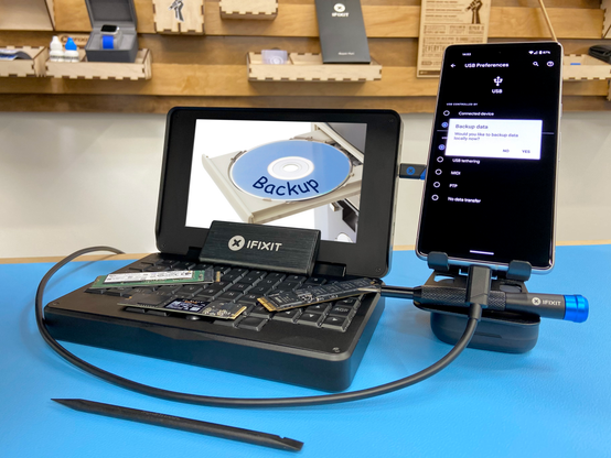 Black MNT Pocket Reform sitting on a blue work mat in front of a wall with wooden french cleats with various items and shelves mounted to it. The laptop has three M.2 2280 SSDs on its keyboard, and one iFixit external M.2 SSD enclosure. The laptop's display shows an image of an older desktop with a beige disc drive. The drive is ejected, revealing a disc with a blue label that reads "Backup" in Comic Sans. Connected to the laptop is a Pixel 7, which is sitting next to it on a phone stand. The phone's display shows the Android USB settings and a dialog box that says "Backup data. Would you like to backup data locally now?" with "NO" and "YES" options. Beneath the phone is an iFixit screwdriver, and in front of the laptop is a lone spudger.