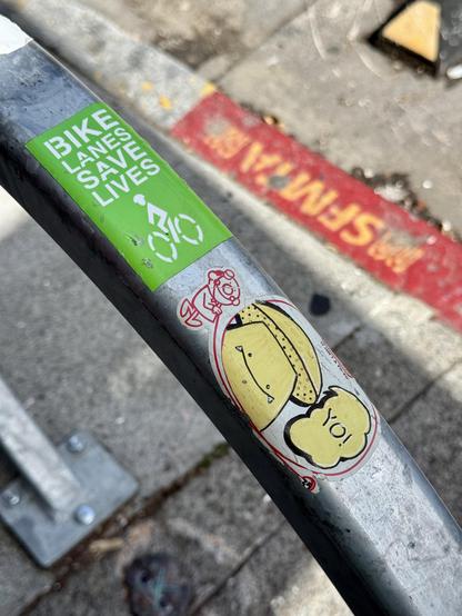 A bike rack with two stickers on it. One is an old Duncan Yo-Yo sticker with graffiti drawn on it depicting a yellow mushroom. The mushroom is drawn in Matsu's iconic style and is saying "Yo!". The other sticker says "Bike Lanes Save Lives". The photo is taken on Valencia Street in San Francisco on a sunny day.