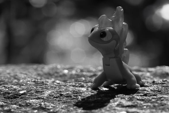Motiv:
Plastikfigur auf einer strukturierten Steinoberfläche. Die Figur steht leicht nach rechts gewendet im Vordergrund und ist von hinten beleuchtet.

Stimmung & Wirkung:
Das Bild vermittelt eine ruhige, isolierte Szene. Die Lichtführung hebt die Figur deutlich vom Hintergrund ab und erzeugt eine klare Silhouette. Die starke Unschärfe im Hintergrund führt zur Konzentration auf das Objekt und erzeugt eine minimalistische Wirkung.

Technische Details:
Farbe: Schwarzweiß. Helle Grautöne dominieren den Hintergrund, dunkle bis mittlere Grautöne bilden den Vordergrund und die Figur.
Schärfentiefe: Gering. Nur die Figur und der direkt angrenzende Untergrund sind scharf, der Hintergrund ist vollständig unscharf mit ausgeprägtem Bokeh.
Lichtverhältnisse: Hartes, gerichtetes Licht von hinten. Klare Lichtkante an der Figur. Der Hintergrund enthält viele helle Lichtreflexe.