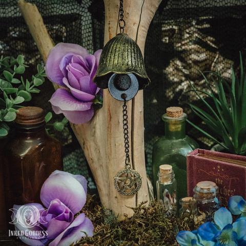 A Moon Phases Wind Chime hangs from a bare wooden branch. It consists of a brass tulip-shaped bell and a black crescent moon clapper with a brass pentacle hanging from the clapper on a black chain. This is an exclusive product from Inked Goddess Creations. It was included in our February 2025 "Witchy Shit 3" IGC Box.