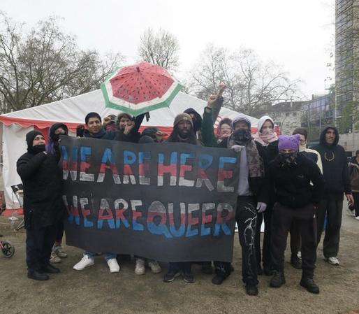 A group of humans standing at oplatz in front of a big tent, with a banner, that reads "we are here, we are queer", written in the colors of the trans flag. There is also an umbrella with watermelon design.