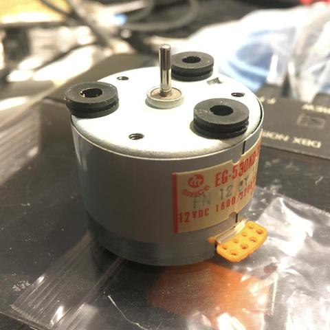 An EG-530KD-2B capstan motor, freshly removed from a Tascam 424mkII multitrack cassette recorder, sits on its back on top of a plastic bag, which itself is on top of a Tascam Porta05 machine. There are three black rubber vibration grommets resting on top of it.