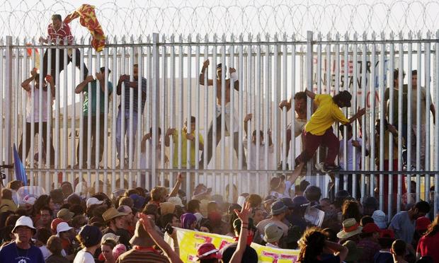 Photograph of the white steel fence at Woomera Detention centre some refugees are climbing over and through the bars.

text:
1,000 activists rallied outside the detention centre on 30 March 2002, the refugees held their own protest inside the centre, and when the demonstrators outside approached, they began scaling the five-metre-high fence and leaping into the crowd.
Although most of the 50 refugees who escaped that day were subsequently captured and returned to imprisonment, some were never caught. With ongoing protests from refugees at Woomera, many were granted permanent visas - including the 13 who were not recaptured on 30 March - and by 2003 the centre was permanently closed. While the Australian government's treatment of refugees has subsequently worsened, refugees' actions at Woomera in 2002 showed that militant protest could force governments to grant concessions and could begin to turn the tide of mass opinion.