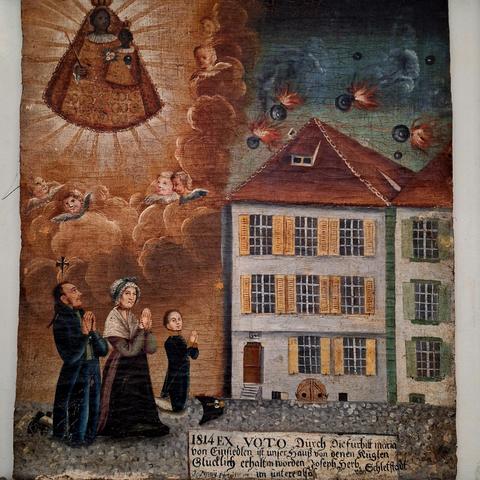 The painting depicts a couple and a child on their knees, praying towards a three-story building. Above the building, flaming cannonballs are visible. Above the family, the Black Madonna of Einsiedeln is shown floating in the sky. The painting was commissioned by Joseph Herb from Schletstadt im Unterelsass, a city now called Sélestat in France. The bombing likely refers to the siege by Bavarian troops in 1814, during which the city did not fall, although it suffered several hundred deaths and an epidemic of typhus.