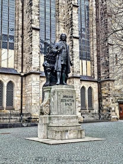 Das Bild zeigt die Statue von Johann Sebastian Bach vor der Thomaskirche in Leipzig. Die Bronzestatue stellt den berühmten Komponisten des Barock in typischer Kleidung seiner Zeit dar: ein langer Mantel, Kniehosen und eine Perücke. In seiner linken Hand hält er eine Schriftrolle, während seine rechte Hand auf eine kunstvoll verzierte Orgel zeigt, die sich neben ihm befindet. Der steinerne Sockel der Statue trägt die Inschrift „JOHANN SEBASTIAN BACH“.

Die Kulisse bildet die Fassade der Thomaskirche mit ihren hohen gotischen Fenstern und der detailreichen Steinarchitektur. Der gepflasterte Platz vor der Statue ist weitgehend leer, nur einige kahle Äste eines Baumes ragen am rechten Bildrand ins Bild, was auf eine winterliche oder frühe Frühlingszeit hindeutet.