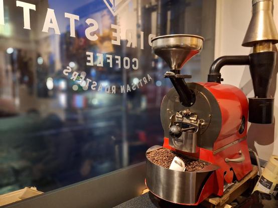 Picture taken from the inside of a shop onto a dark street. There is a shop name printed on the window, which mirrored reads "[Emp]ire Stat[e] Coffee - Artisan Roasters". On the right hand side of the picture sits a red coffee roasting machine.