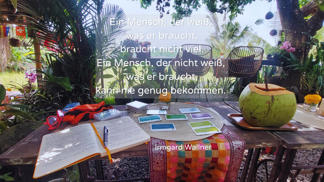 Ein stiller Moment auf Bali: Karten, Buch & Kokosnuss auf einem Holztisch mit Blick ins Grüne. Zitat über Bedürfnisse eingeblendet