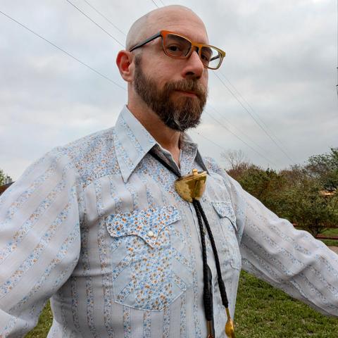 A photo of me, a 47 year old white man with a shaved head, a short cropped brown & grey beard, 

photographed from the chest up

Against a grey cloudy sky and trees in a park

wearing

rectangular pearlescent orange acetate glasses, a small brass semicircular bowl with a lid and knob as a bolo tie, on a floral blue, white, and orange western shirt 