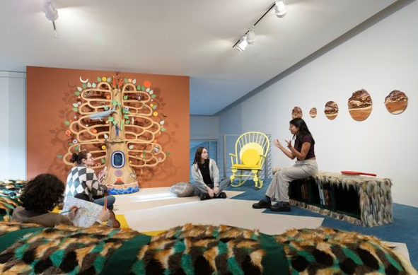 Photo of a white gallery room with people sitting on comfortable soft benches and talking to each other. One person is reading? sketching? On one wall is a symbolic tree with bright leaves. Another wall has several round pieces like desert sand dunes.