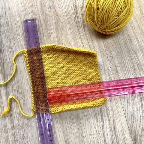 A yellow knitted swatch in stockinette stitch is shown on a light wooden background. The ball of yarn and 2 rulers are also visible.