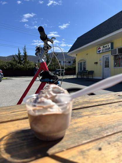 a cup of ice cream on a table, my red bike behind it. In the distance Ron’s Ice Cream is seen, and in the far distance the hills behind our town. 