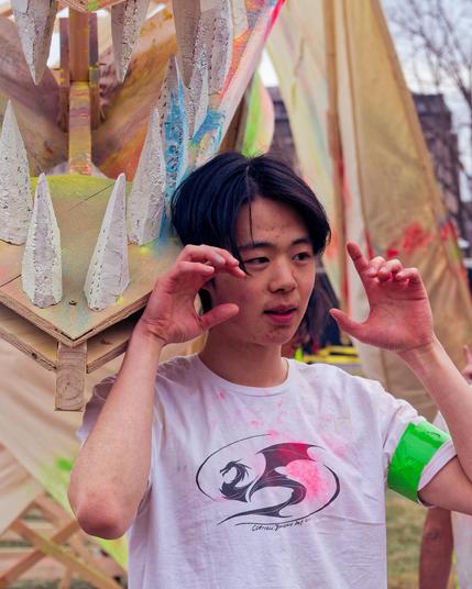 Asian man wearing a white t-shirt with a dragon logo splattered with red paint and with a green stripe of tape on his arm poses with his hands up like claws in front of a wood-framed dragon float with numerous sharp teeth