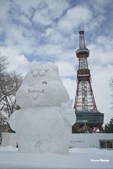 A photograph from the Sapporo Snow Festival 2025.  The foreground sees a cheeky little mascot sculpted in snow, tongue out and fingers held in a peace sign.  In the background stands Sapporo TV tower.