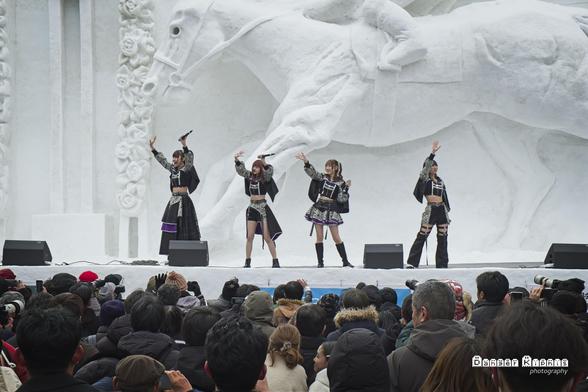 A photograph from the Sapporo Snow Festival 2025.  4 singing idols in black outfits with silver details wave to the crowd while performing on a stage sculpted out of snow.  The stage backdrop is a large snow sculpture of a racehorse and jockey.