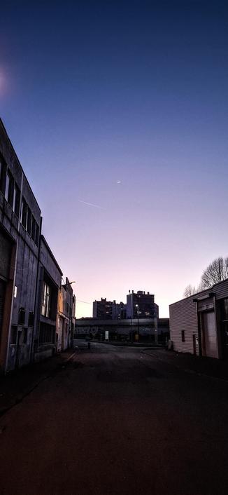 Vue d'un faubourg de saint Étienne, avec un ciel de Crépuscule en chien et loup. On peut voir des bâtiments industriels de part et d'autre de la rue, et deux bâtiments d'habitation derrière la voie rapide. Un petit croissant de lune 🌙  est visible dans le ciel ainsi qu'un trait de turbo réacteur d un avion.