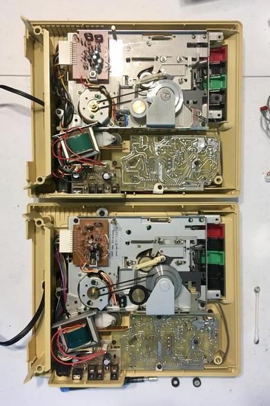 Two yellow Library of Congress C-1 cassette players rest on a white table, face down, with their back covers removed. This has exposed various internal PCBs, the motor and the rear of the cassette mechanism, with belts and pulleys visible.
