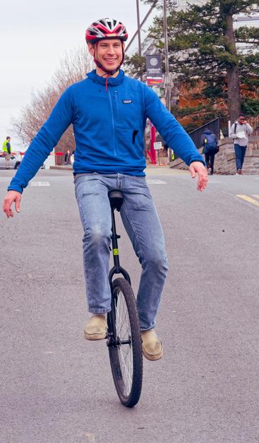 A man in a blue jacket and wearing blue jeans and a red and silver bike helmet rides on a black unicycle on the road