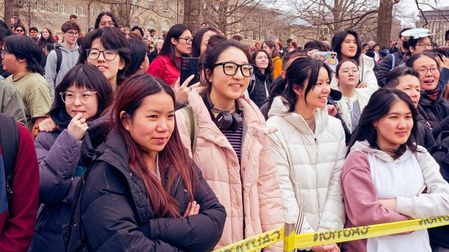 Four young asian women are in the front row of a large crowd of spectators underneath a canopy of  winter phase trees and with the stone and window facade of Goldwin Smith Hall in the background and caution tape crossing the lower right corner.