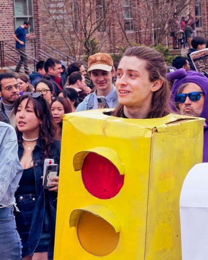 A man in a traffic light costume has visible red and yellow light panels and is surrounded by a dense crowd of other parade goers.