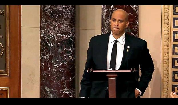 Senator Cory Booker stands at a podium in the U.S. Senate chamber, wearing a black suit and tie with a small American flag and gold ribbon pinned to his lapel. He has a serious expression, with one hand in his pocket, framed by marble columns and ornate architectural details.