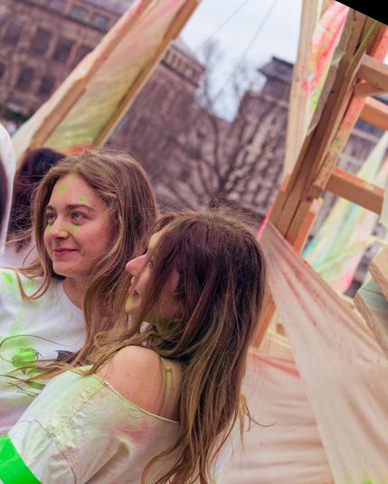 Two girls splattered with green paint pose against a wooden frame with stretched fabric panels in an image that you can see has been rotated because buildings rise up diagonally in the background