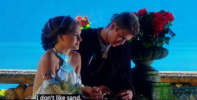 Scene from Days of Our Lives, a US daytime soap opera. (JK it's Star Wars)  Scene is on a Oceanview balcony with vast blue water in the distance and blue sky. Closer in we see a princess type girl with hair jangles and fancy neck ware and shimmery dress, standing next to an angsty looking emo boy in all black. He has one long strand of hair with an otherwise short cut, like a hair bolo or something. Behind them is a big patio bouquet of red flowers. She's looking happy and dreamy, he's looking down and probably wishing he was listening to Korn and smoking cloves. Caption reads, "I don't like sand."