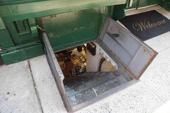 Open metal access hatch to a below-street-level space, featuring interior stairs and food preparation area.